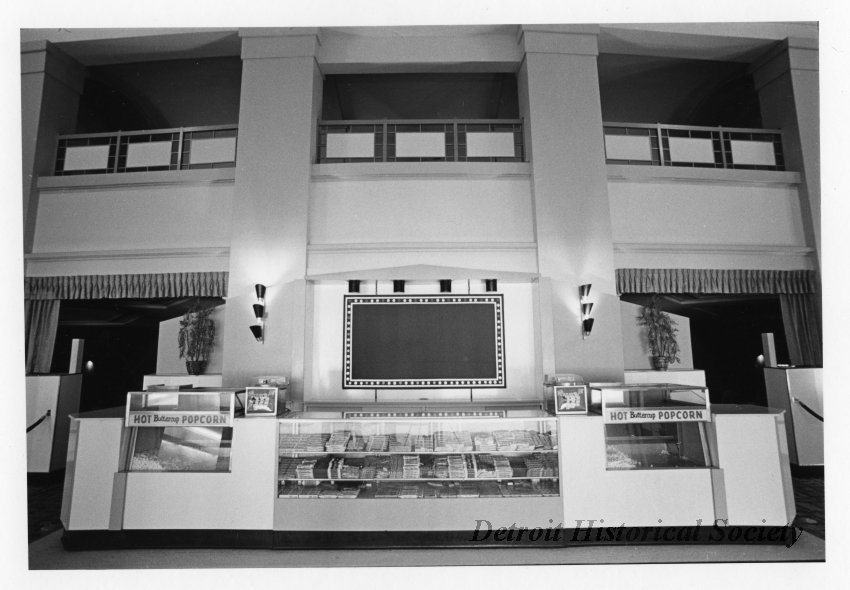 Black and white photographic print depicting a concession stand in the lobby of the Redford Theatre. The mezzanine is visible overhead. Signs on counter read "Hot Buttercup Popcorn".