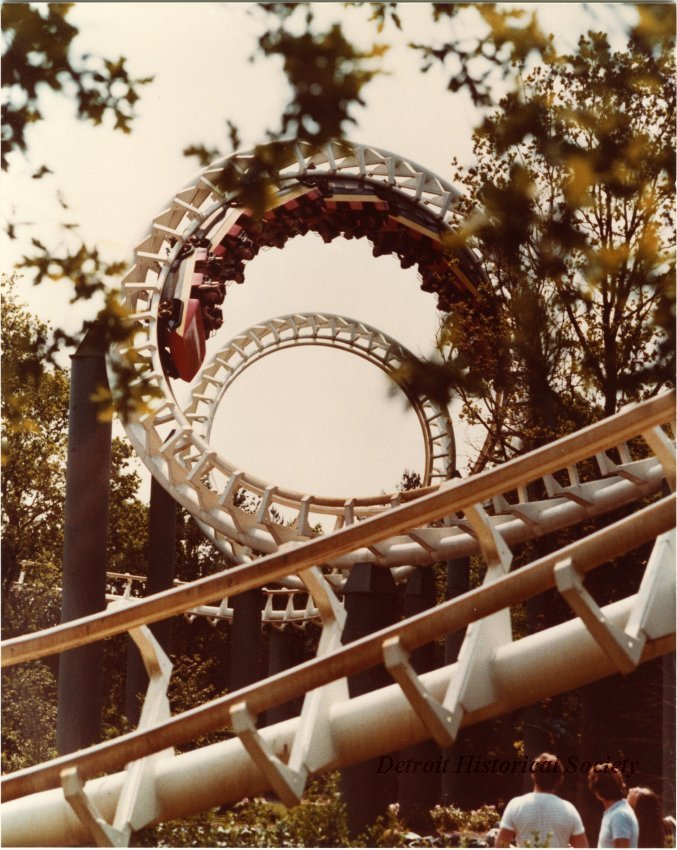 Color photographic print taken of the double corkscrew portion of Boblo Island's roller coaster, the Screamer. The ride's train is inverted in the second corkscrew. Several people watch from the ground in the lower right corner.