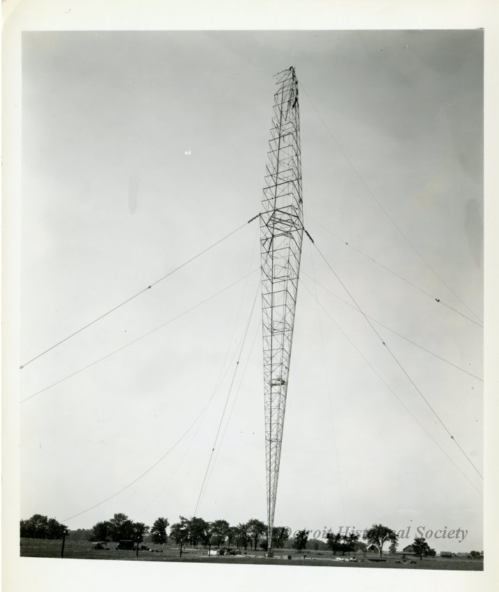 Black and white photo of the construction of the WJR antenna outside of the WJR Radio Transmitting Building on the southwest corner of Sibley Road and Grange Road in Riverview. Several construction workers and piles of building material are scattered around the base of the tower.