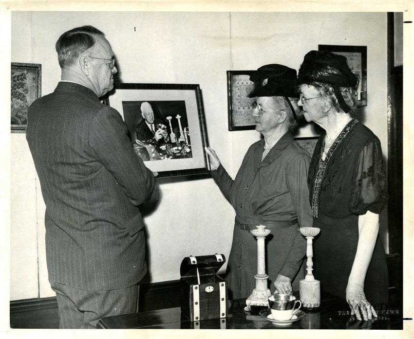 Black and white photographic print of (left to right) J. Bell Moran; Gracie B. Krum; and Mrs. Arthur S. Hampton, viewing photographs, and artifacts inside the Detroit Historical Museum, 1949.