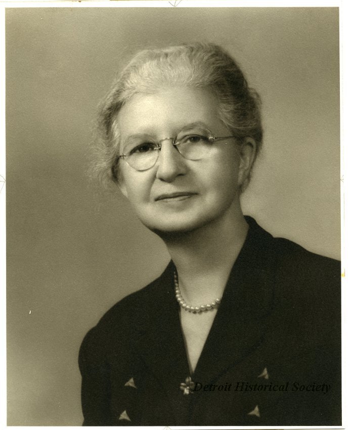 Sepia-toned head-and-shoulders portrait photograph of Gracie Krum. She is wearing a suit jacket with a vertical triangle design on each side of her jacket, and a flower where her lapels meet. She also wears a pearl necklace, her hair pulled back, and glasses.