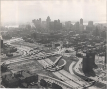 Black and white aerial photograph depicting a view along I-75, looking south from the Division Street pedestrian bridge, with the I-375 interchange under construction. In the lower right is the Brewster-Douglass Housing Projects. In the lower left is Bishop Union School. In the background is the downtown skyline. 