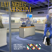 A museum exhibit featuring sports memorabilia from the Detroit Lions, including jerseys, helmets, and a life-size football player cutout in a glass-enclosed space.