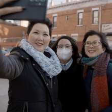 Three Aisian women taking a selfie. The woman holding the camera is wearing a black coat and a grey infinity scarf. The woman in the center of the group is wearing a mask. The third woman is wearing classes and a red and maroon scarf.