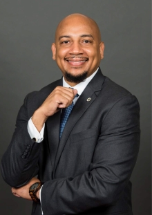 Dr. Ken L. Harris, a smiling Black man in a suit and tie posing with his hand on his chin against a gray background.