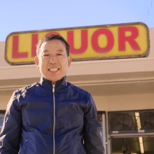 Man in a blue zip up jacket standing in front of a store front with a liquor sign. His head is blocking the letter Q.