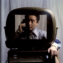 Man holding a corded phone receiver to his ear, viewed through an old-fashioned television screen.