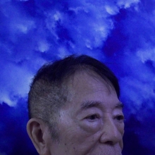 Top half of an older Asian man's face on a dark blue cloudy background.