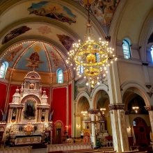 Interior of ornate catholic church with lighted crystal chandelier hanging from the painted ceiling and an altar in front of a red wall.