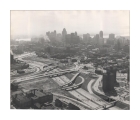 Black and white aerial photograph depicting a view along I-75, looking south from the Division Street pedestrian bridge, with the I-375 interchange under construction. In the lower right is the Brewster-Douglass Housing Projects. In the lower left is Bishop Union School. In the background is the downtown skyline. 